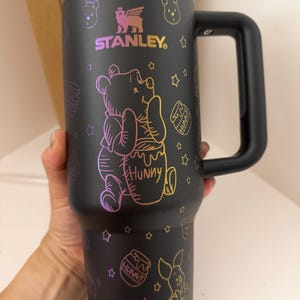 Puede incluir: Un vaso Stanley negro mate con asa y borde iridiscente. El exterior presenta un colorido patrón de líneas de estilo holográfico de Winnie the Pooh, Piglet y tarros de miel etiquetados como "Hunny", intercalados con pequeñas formas de estrellas.