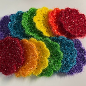 May include: A set of rainbow colored crocheted scrubbers with a sparkly finish. The scrubbers are arranged in a semi-circle, with the colors transitioning from purple to red, orange, yellow, green, blue, and purple.