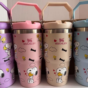May include: Four Stanley tumblers in pastel colors: purple, pink, beige, and blue. Each tumbler features a stainless steel interior, a matching handle and lid, and a cartoon character design with hearts, clouds, and bones. The Stanley logo is visible on each tumbler.