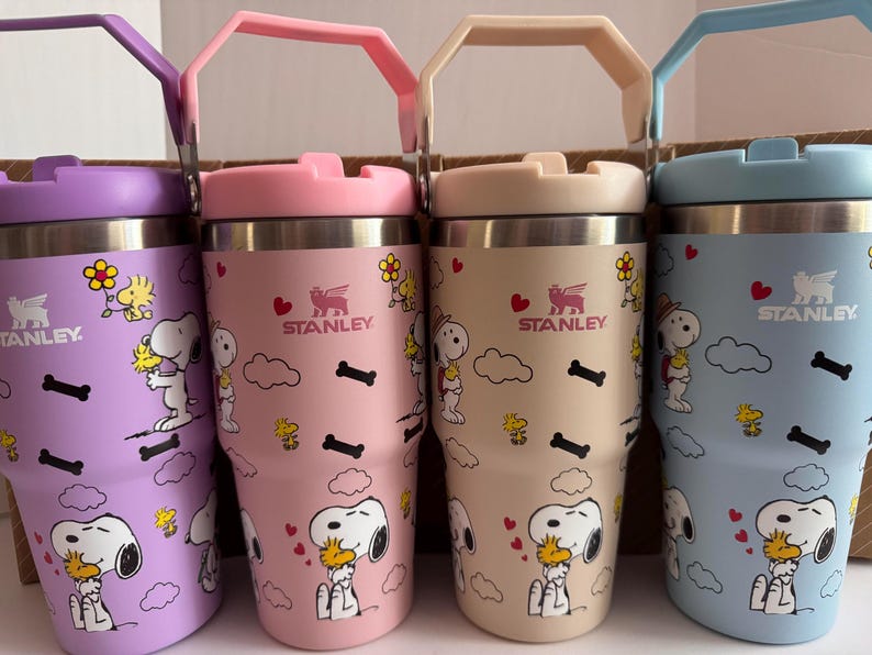 May include: Four Stanley tumblers in pastel colors: purple, pink, beige, and blue. Each tumbler features a stainless steel interior, a matching handle and lid, and a cartoon character design with hearts, clouds, and bones. The Stanley logo is visible on each tumbler.