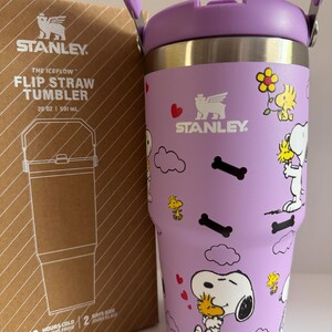 May include: A purple Stanley The Iceflow Flip Straw Tumbler, 20 oz, featuring Snoopy and Woodstock graphics. The tumbler has a purple handle and lid, with the Stanley logo. The box next to it has the Stanley logo and product information.