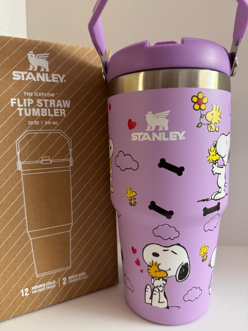 May include: A purple Stanley The Iceflow Flip Straw Tumbler, 20 oz, featuring Snoopy and Woodstock graphics. The tumbler has a purple handle and lid, with the Stanley logo. The box next to it has the Stanley logo and product information.