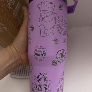 Puede incluir: Un vaso t&eacute;rmico de color lavanda con Winnie the Pooh y sus amigos. El vaso tiene un asa y tapa moradas, con un acento plateado. El dise&ntilde;o incluye ilustraciones de Winnie the Pooh, Tigger, abejas, flores y un tarro de miel.