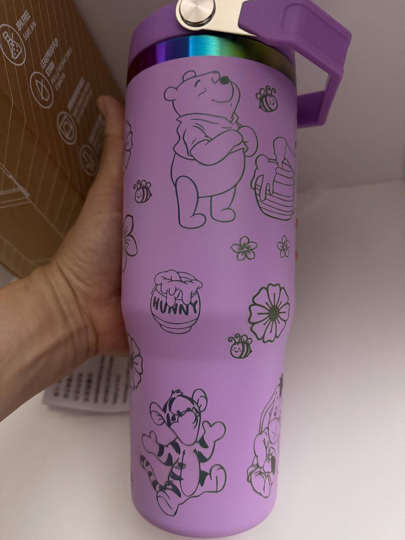 Puede incluir: Un vaso t&eacute;rmico de color lavanda con Winnie the Pooh y sus amigos. El vaso tiene un asa y tapa moradas, con un acento plateado. El dise&ntilde;o incluye ilustraciones de Winnie the Pooh, Tigger, abejas, flores y un tarro de miel.