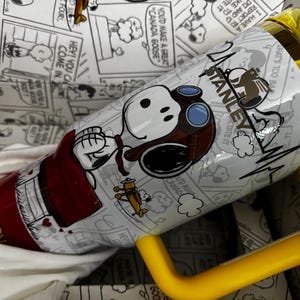 May include: A white Stanley tumbler featuring a graphic of Snoopy wearing aviator goggles and a brown cap. The design includes a small yellow airplane, the text "SNOOPY" in yellow, and a yellow handle and lid. The background pattern shows black and white comic strips.