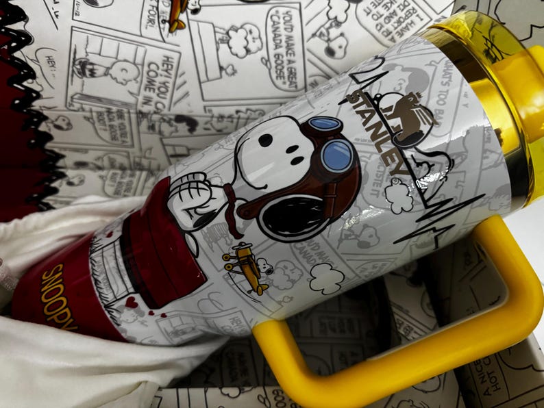 May include: A white Stanley tumbler featuring a graphic of Snoopy wearing aviator goggles and a brown cap. The design includes a small yellow airplane, the text "SNOOPY" in yellow, and a yellow handle and lid. The background pattern shows black and white comic strips.