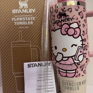 May include: A pink and white Stanley Flowstate tumbler with a Hello Kitty design, a clear straw, and a product box. The tumbler has a capacity of 40 oz (1.18 L). The design features a pink leopard print and a Hello Kitty graphic.