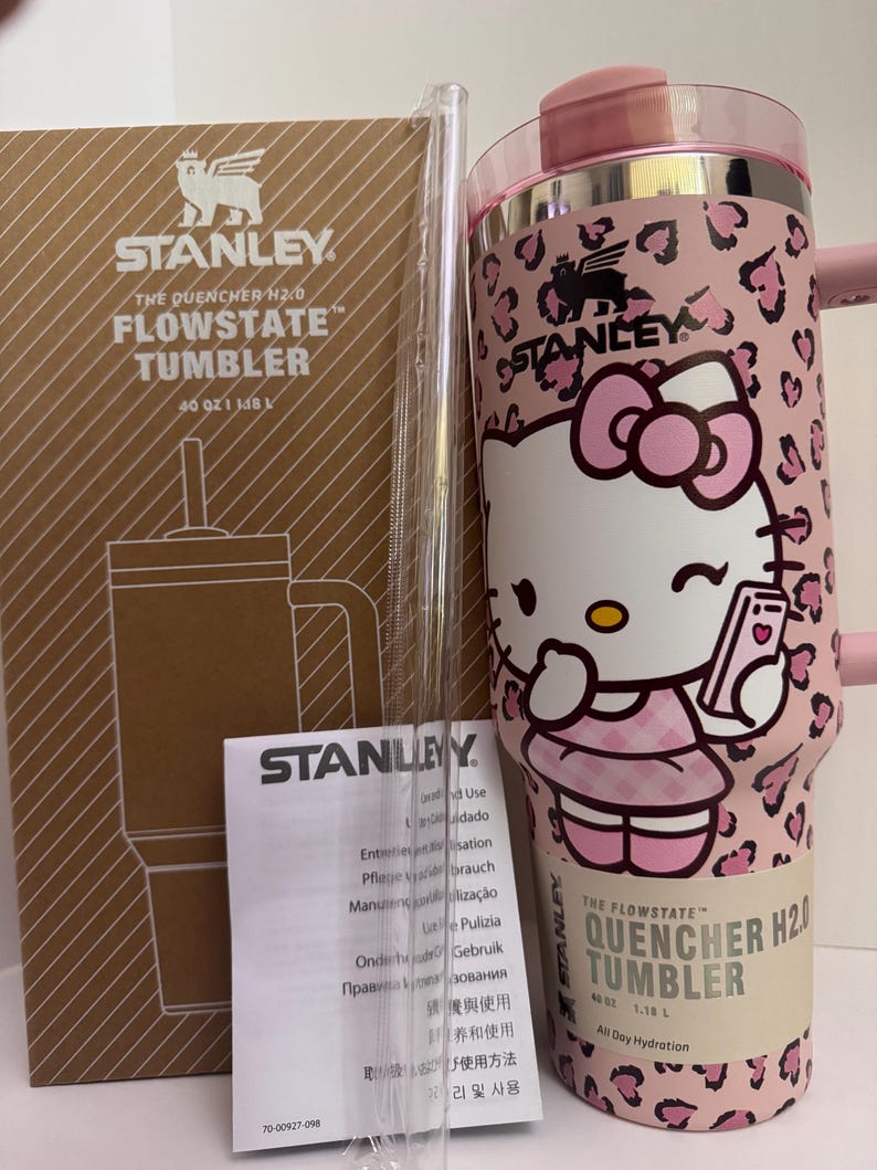 May include: A pink and white Stanley Flowstate tumbler with a Hello Kitty design, a clear straw, and a product box. The tumbler has a capacity of 40 oz (1.18 L). The design features a pink leopard print and a Hello Kitty graphic.