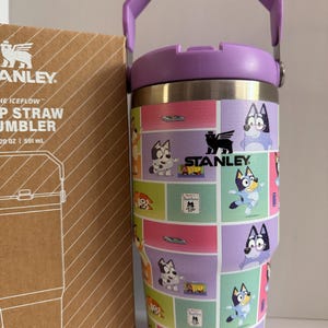 May include: A stainless steel Stanley Iceflow tumbler with a purple handle and lid. The tumbler features a colorful cartoon design with various characters. The box next to it has the Stanley logo and product information.