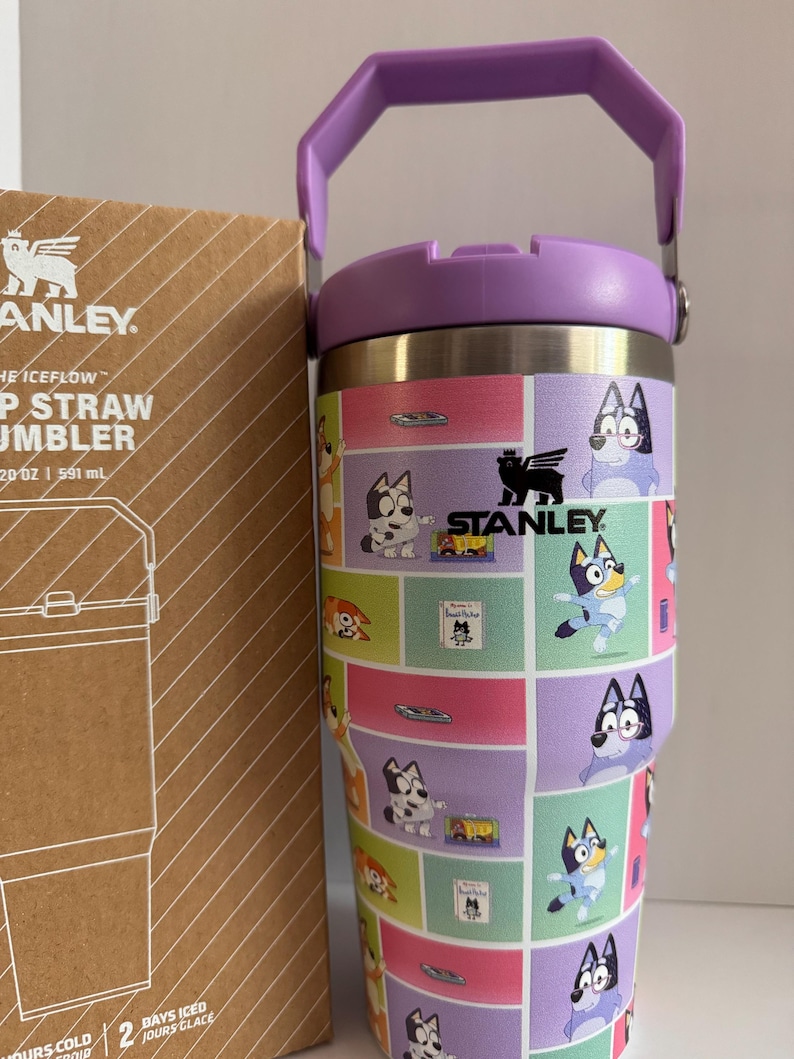 May include: A stainless steel Stanley Iceflow tumbler with a purple handle and lid. The tumbler features a colorful cartoon design with various characters. The box next to it has the Stanley logo and product information.