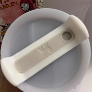 Puede incluir: Tapa de vaso Stanley blanca y gris claro con un deslizador blanco. La tapa tiene un interior gris claro y un emblema beige. El fondo incluye una vista parcial de un vaso con un dise&ntilde;o de Sally.