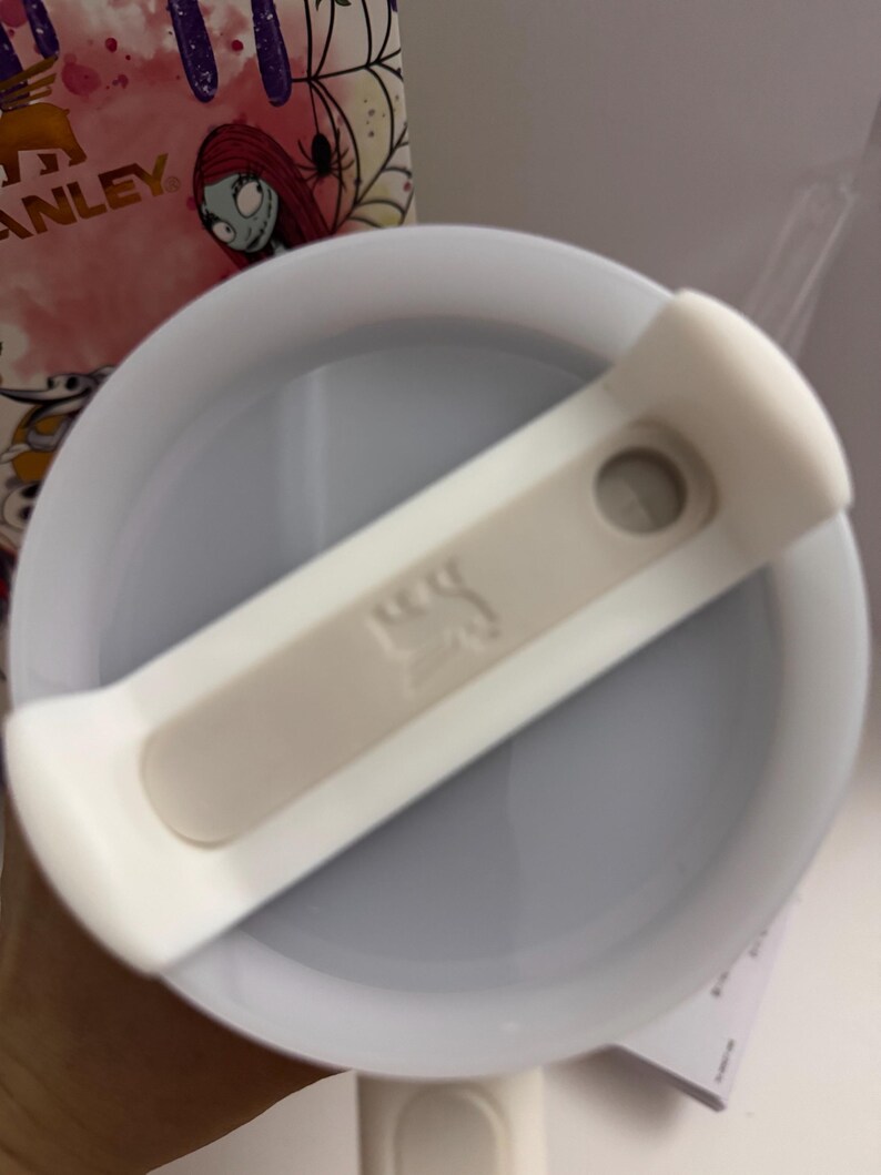 Puede incluir: Tapa de vaso Stanley blanca y gris claro con un deslizador blanco. La tapa tiene un interior gris claro y un emblema beige. El fondo incluye una vista parcial de un vaso con un dise&ntilde;o de Sally.