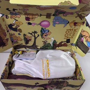 K&ouml;nnte beinhalten: Ge&ouml;ffnete Box mit Winnie-the-Pooh-Motiv. Innenraum und Deckel sind mit Cartoon-Illustrationen von Puuh und seinen Freunden verziert. Eine wei&szlig;e Tasche mit dem Text "LOVESHACKFANCY X STANLEY" befindet sich in der Box.