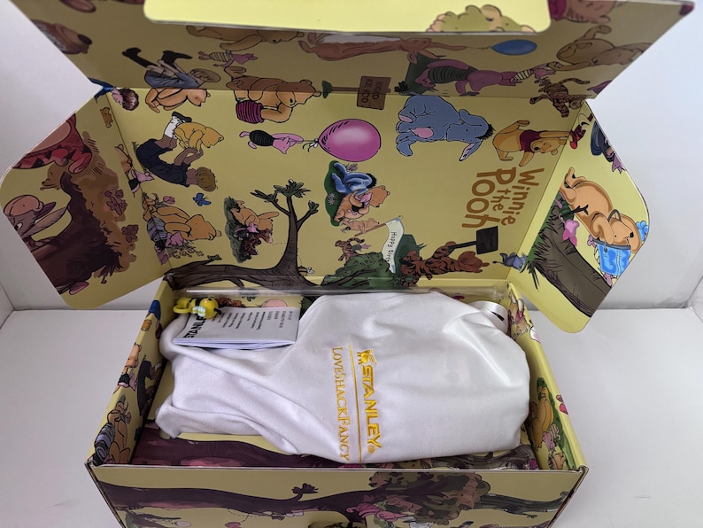K&ouml;nnte beinhalten: Ge&ouml;ffnete Box mit Winnie-the-Pooh-Motiv. Innenraum und Deckel sind mit Cartoon-Illustrationen von Puuh und seinen Freunden verziert. Eine wei&szlig;e Tasche mit dem Text "LOVESHACKFANCY X STANLEY" befindet sich in der Box.