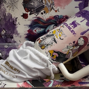 Puede incluir: Una taza Stanley con personajes de Pesadilla antes de Navidad, con un asa blanca y una bolsa con cord&oacute;n blanca. La taza tiene un fondo rosa con detalles morados e ilustraciones de personajes. El interior de la caja tiene ilustraciones a juego.