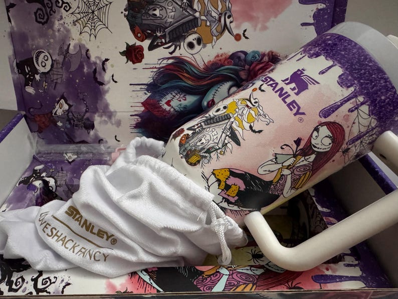Puede incluir: Una taza Stanley con personajes de Pesadilla antes de Navidad, con un asa blanca y una bolsa con cord&oacute;n blanca. La taza tiene un fondo rosa con detalles morados e ilustraciones de personajes. El interior de la caja tiene ilustraciones a juego.