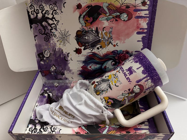 Puede incluir: Un vaso Stanley Loveshack Fancy con asa blanca y tapa blanca, dentro de una caja decorativa. El vaso presenta una colorida ilustraci&oacute;n de personajes. La caja y el vaso tienen una combinaci&oacute;n de colores p&uacute;rpura y rosa.