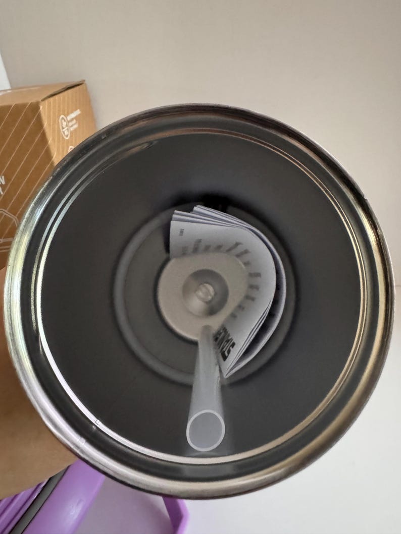 May include: A stainless steel tumbler with a gray interior, a clear straw, and a lid with a central opening. The lid has a small, circular opening for the straw. A folded paper with text is inside the tumbler.
