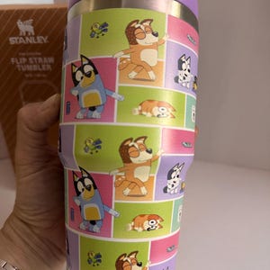 May include: A Stanley Flip Straw Tumbler with a purple lid and a colorful wrap featuring cartoon dog characters in various poses. The tumbler has a stainless steel body and a flip-top straw.