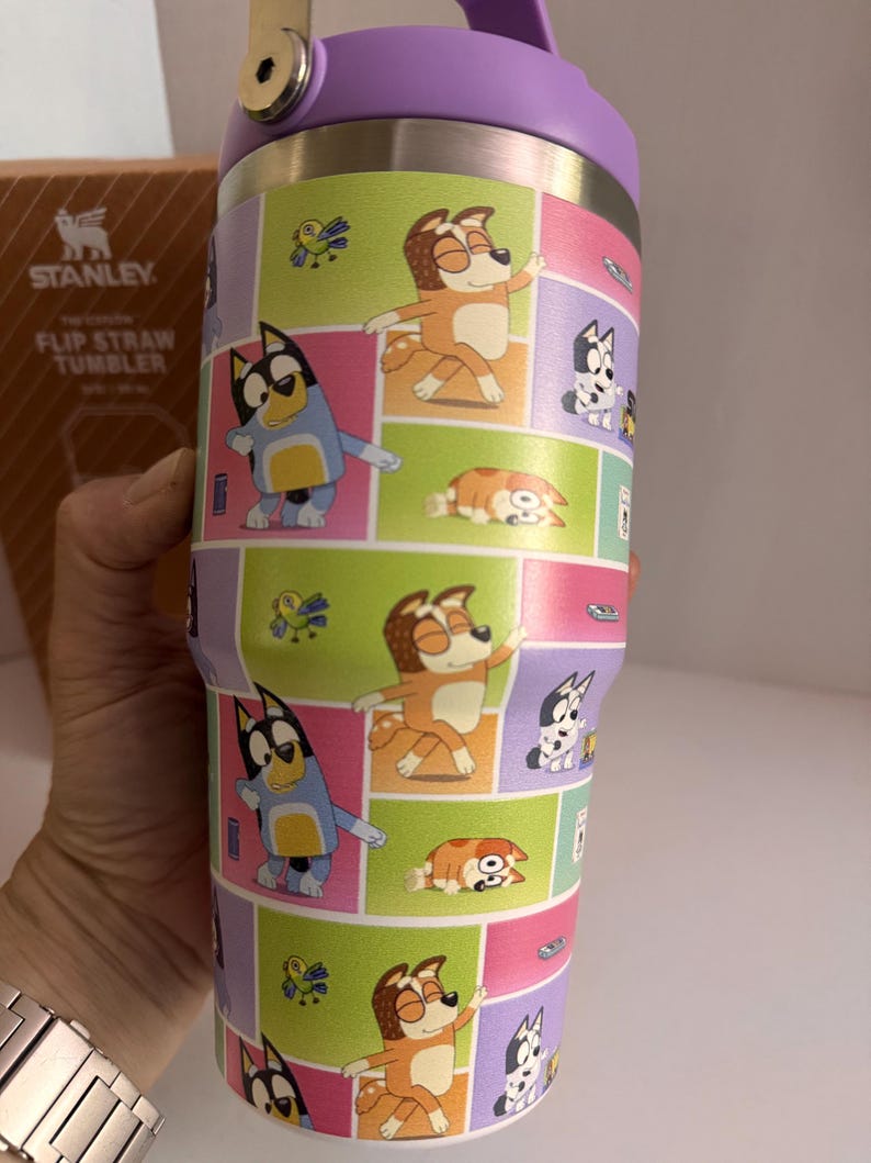 May include: A Stanley Flip Straw Tumbler with a purple lid and a colorful wrap featuring cartoon dog characters in various poses. The tumbler has a stainless steel body and a flip-top straw.