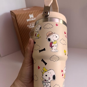 May include: A beige Stanley Flip Straw Tumbler featuring Snoopy and Woodstock cartoon graphics. The tumbler has a cream-colored handle and lid, with a silver metal clasp. The design includes Snoopy in various poses, clouds, hearts, and black bone shapes.