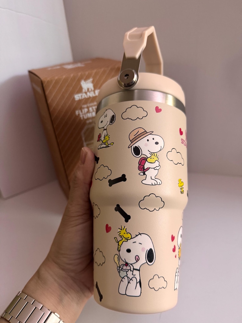 May include: A beige Stanley Flip Straw Tumbler featuring Snoopy and Woodstock cartoon graphics. The tumbler has a cream-colored handle and lid, with a silver metal clasp. The design includes Snoopy in various poses, clouds, hearts, and black bone shapes.