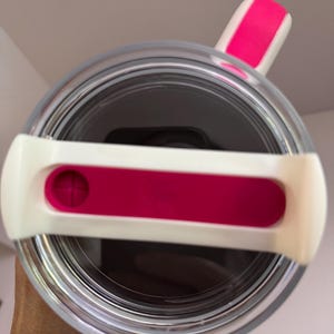 Op de afbeelding: Een heldere, cilindrische beker met een wit en roze deksel. Het deksel heeft een roze schuif en een witte handgreep. De binnenkant van de beker lijkt donker en het ontwerp wordt van bovenaf bekeken.