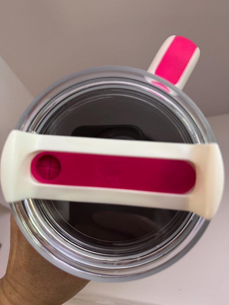 Op de afbeelding: Een heldere, cilindrische beker met een wit en roze deksel. Het deksel heeft een roze schuif en een witte handgreep. De binnenkant van de beker lijkt donker en het ontwerp wordt van bovenaf bekeken.