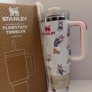 Op de afbeelding: Een witte Stanley Flowstate-beker met een Daisy Duck-patroon, een roze handvat en een doorzichtig rietje. De beker staat naast de originele kartonnen doos, met het Stanley-logo en productinformatie.