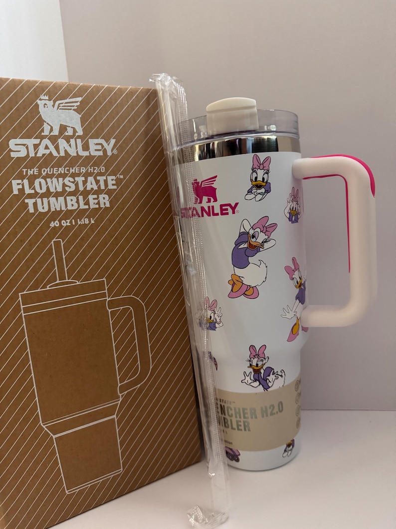 Op de afbeelding: Een witte Stanley Flowstate-beker met een Daisy Duck-patroon, een roze handvat en een doorzichtig rietje. De beker staat naast de originele kartonnen doos, met het Stanley-logo en productinformatie.