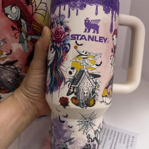 Puede incluir: Un vaso Stanley blanco grande con tapa y asa moradas. El vaso est&aacute; decorado con personajes e im&aacute;genes de Pesadilla antes de Navidad, incluyendo a Sally, Jack Skellington y murci&eacute;lagos. El logotipo de Stanley es visible.