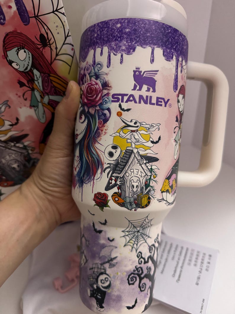 Puede incluir: Un vaso Stanley blanco grande con tapa y asa moradas. El vaso est&aacute; decorado con personajes e im&aacute;genes de Pesadilla antes de Navidad, incluyendo a Sally, Jack Skellington y murci&eacute;lagos. El logotipo de Stanley es visible.