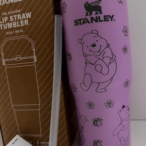 Puede incluir: Un vaso con pajita Stanley morado con un dise&ntilde;o de Winnie the Pooh. El vaso tiene una capacidad de 887 ml e incluye una pajita transparente. El dise&ntilde;o presenta a Winnie the Pooh, flores, abejas y un conejo. El logotipo de Stanley es visible.