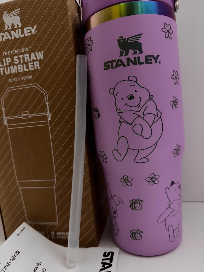 Puede incluir: Un vaso con pajita Stanley morado con un dise&ntilde;o de Winnie the Pooh. El vaso tiene una capacidad de 887 ml e incluye una pajita transparente. El dise&ntilde;o presenta a Winnie the Pooh, flores, abejas y un conejo. El logotipo de Stanley es visible.