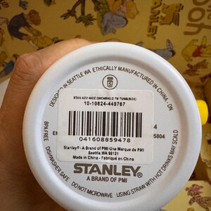 May include: White Stanley tumbler with black text and a barcode. The text includes "STANLEY" and "A BRAND OF PMI". The tumbler is BPA free and dishwasher safe. The background is a yellow pattern.