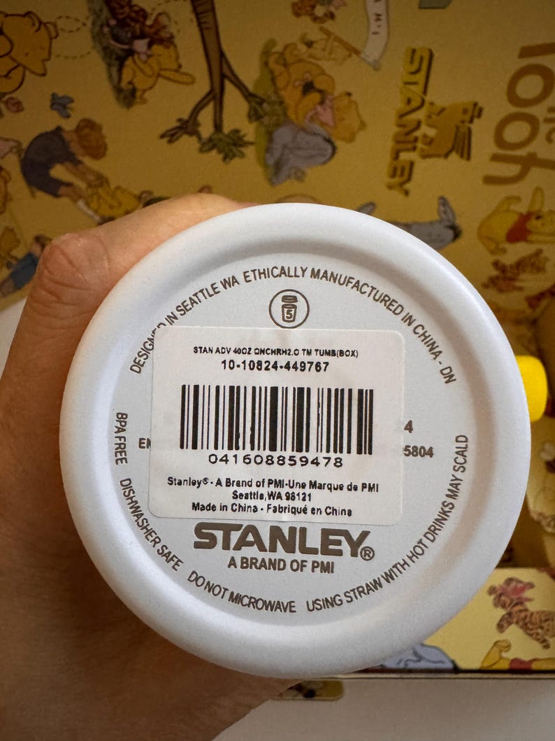 May include: White Stanley tumbler with black text and a barcode. The text includes "STANLEY" and "A BRAND OF PMI". The tumbler is BPA free and dishwasher safe. The background is a yellow pattern.