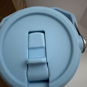 Puede incluir: Primer plano de una tapa de botella de agua azul claro. La tapa tiene un cierre abatible con un bot&oacute;n rectangular y un asa. La tapa est&aacute; hecha de pl&aacute;stico liso y mate. El dise&ntilde;o es simple y funcional.
