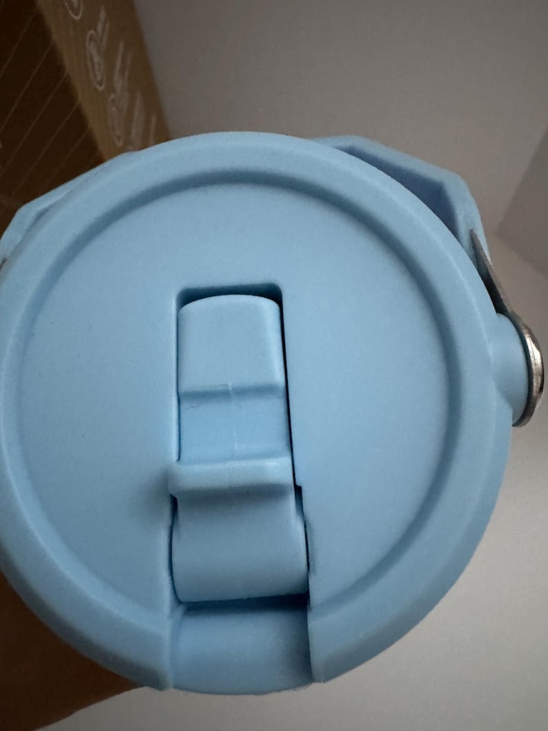 Puede incluir: Primer plano de una tapa de botella de agua azul claro. La tapa tiene un cierre abatible con un bot&oacute;n rectangular y un asa. La tapa est&aacute; hecha de pl&aacute;stico liso y mate. El dise&ntilde;o es simple y funcional.