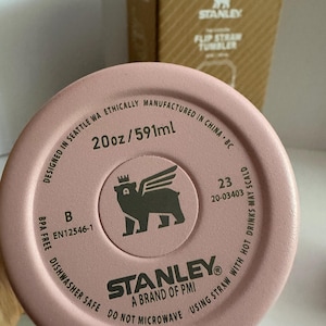 May include: Close-up of a pink Stanley Flip Straw Tumbler lid. The lid features the brand logo, a bear with wings, and text indicating it is 20oz (591ml), BPA-free, and dishwasher safe. A cardboard box is in the background.