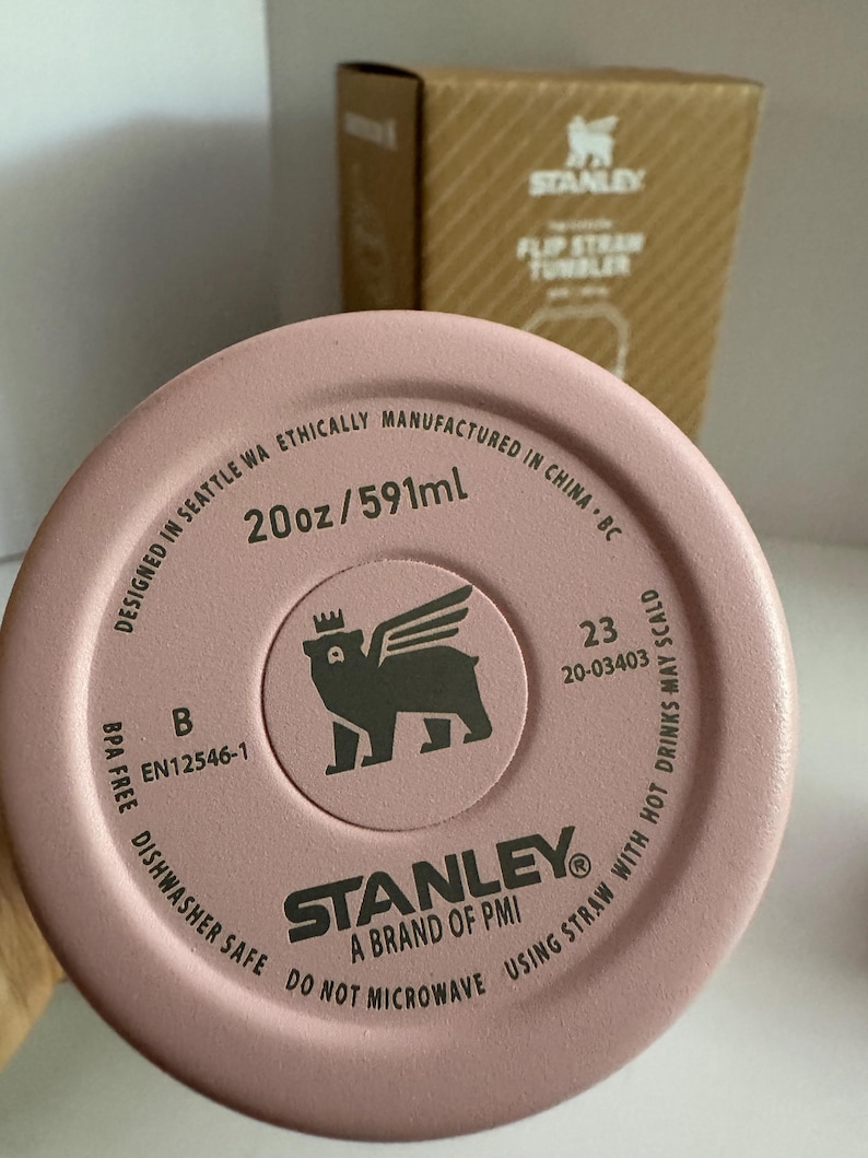 May include: Close-up of a pink Stanley Flip Straw Tumbler lid. The lid features the brand logo, a bear with wings, and text indicating it is 20oz (591ml), BPA-free, and dishwasher safe. A cardboard box is in the background.