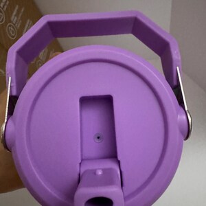 Puede incluir: Vista de cerca de una tapa de botella de agua morada con asa. La tapa tiene una abertura abatible y un peque&ntilde;o respiradero. El asa tiene una forma geom&eacute;trica robusta, y la tapa es de un material liso y s&oacute;lido.