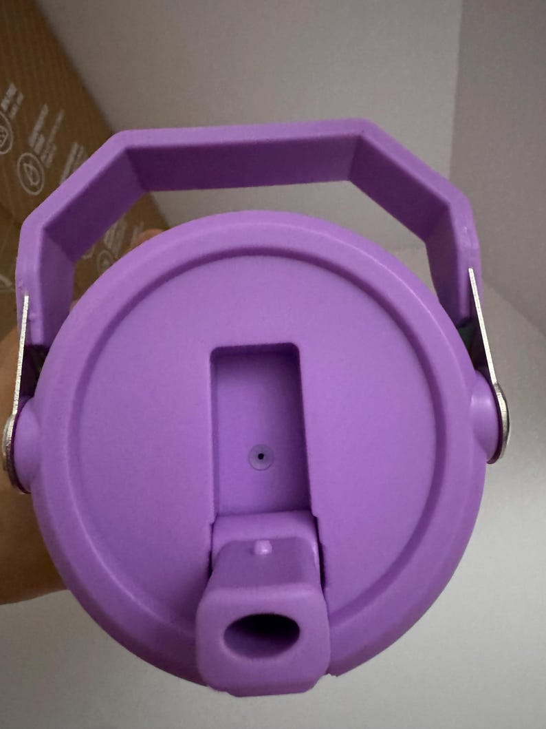 Puede incluir: Vista de cerca de una tapa de botella de agua morada con asa. La tapa tiene una abertura abatible y un peque&ntilde;o respiradero. El asa tiene una forma geom&eacute;trica robusta, y la tapa es de un material liso y s&oacute;lido.