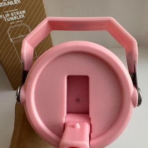 May include: Pink Stanley Flip Straw Tumbler lid with a handle. The lid has a flip-top straw opening and is attached to the tumbler with metal hinges. The box in the background has the Stanley logo and product information.