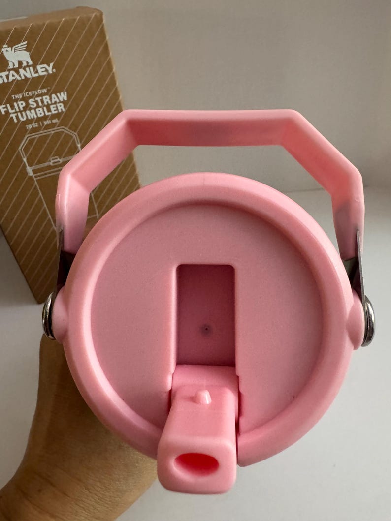 May include: Pink Stanley Flip Straw Tumbler lid with a handle. The lid has a flip-top straw opening and is attached to the tumbler with metal hinges. The box in the background has the Stanley logo and product information.