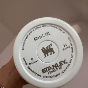 Op de afbeelding: Witte bodem van een Stanley-beker met de tekst "40oz/1.18L". De beker is vaatwasmachinebestendig en BPA-vrij. Het Stanley-logo is zichtbaar, samen met de tekst "Designed in Seattle WA Ethically Manufactured in China-BC".