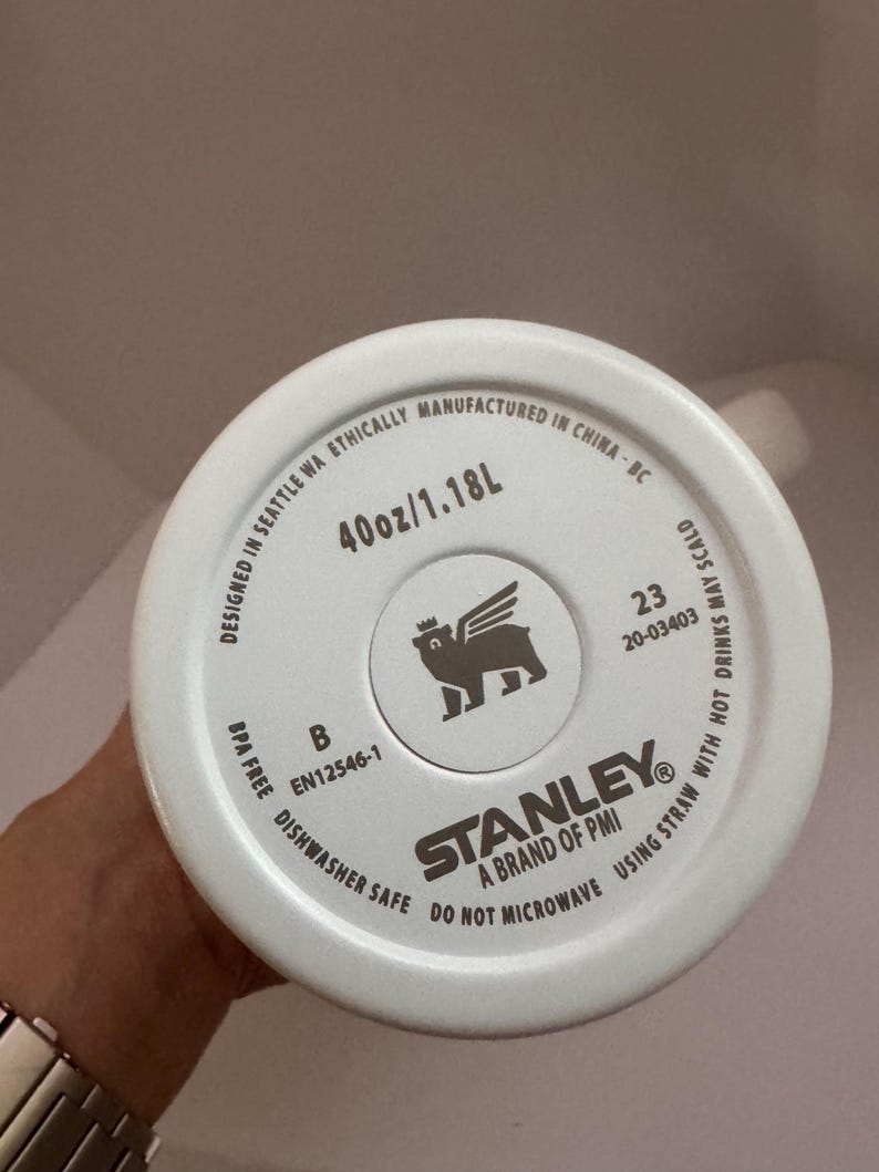 Op de afbeelding: Witte bodem van een Stanley-beker met de tekst "40oz/1.18L". De beker is vaatwasmachinebestendig en BPA-vrij. Het Stanley-logo is zichtbaar, samen met de tekst "Designed in Seattle WA Ethically Manufactured in China-BC".