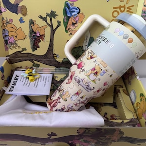 K&ouml;nnte beinhalten: Ein Stanley H2.0 Quencher-Becher mit Winnie-the-Pooh-Design, hellblauem Deckel und Griff. Der Becher befindet sich in einer gelben Box mit Winnie-the-Pooh-Illustrationen. Eine kleine Bienenfigur und ein wei&szlig;es Tuch sind ebenfalls enthalten.