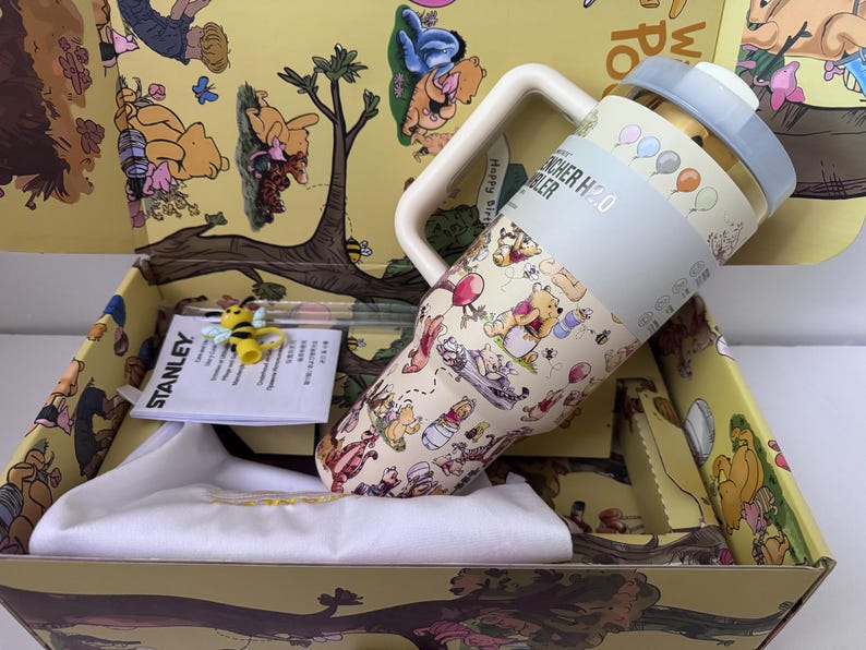 K&ouml;nnte beinhalten: Ein Stanley H2.0 Quencher-Becher mit Winnie-the-Pooh-Design, hellblauem Deckel und Griff. Der Becher befindet sich in einer gelben Box mit Winnie-the-Pooh-Illustrationen. Eine kleine Bienenfigur und ein wei&szlig;es Tuch sind ebenfalls enthalten.