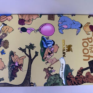 K&ouml;nnte beinhalten: Eine rechteckige Box mit gelbem Hintergrund mit Illustrationen von Winnie-the-Pooh-Charakteren. Die Box hat die Worte "Winnie the Pooh" in brauner Schrift. Die Box hat auch das Stanley-Logo.