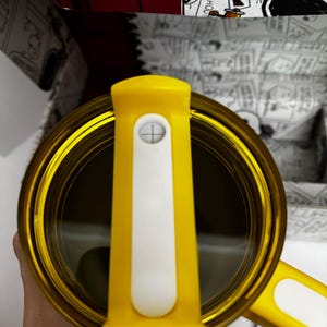 May include: A yellow travel mug with a clear lid and a white sliding closure mechanism. The mug features a yellow handle and is positioned in front of a box decorated with black and white comic strip illustrations of a cartoon dog character.